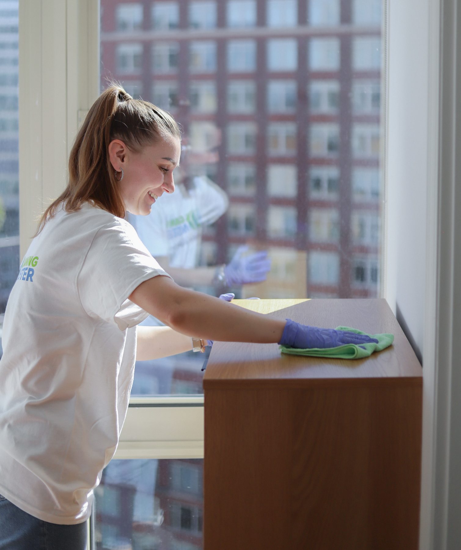 Cleaning Master professional dusting a bedroom dresser beside a New York City window